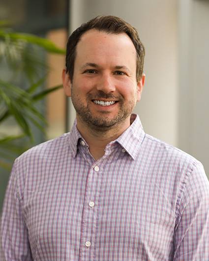 Greg Ducker smiling in a plaid button-up shirt against a blurred background